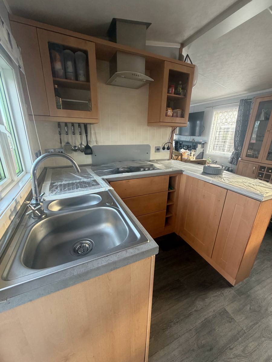 L-shaped caravan kitchen with maple-effect doors and a stainless sink, before